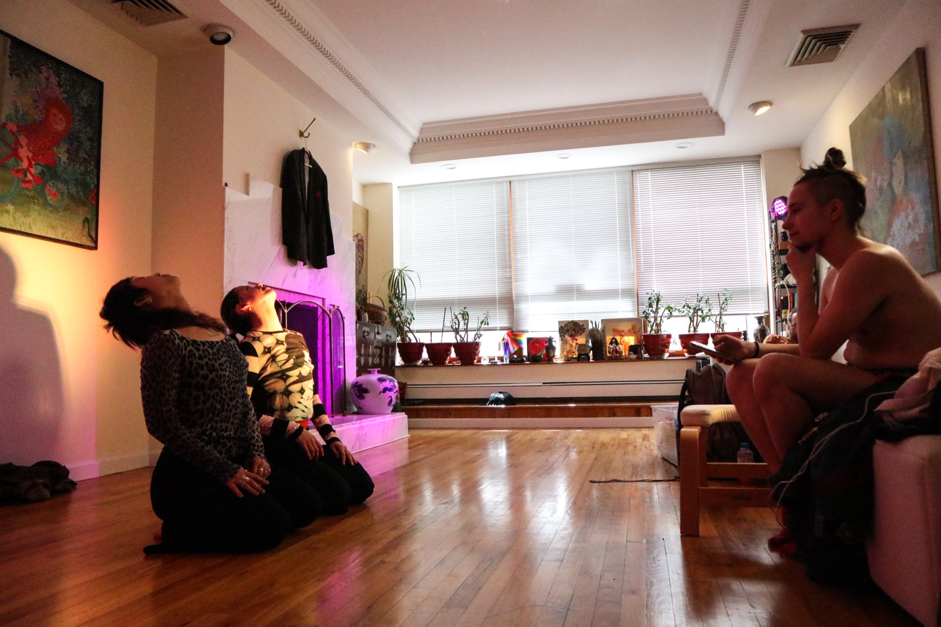 Two performers in the studio, fireplace glowing violet — a witness watches from the side